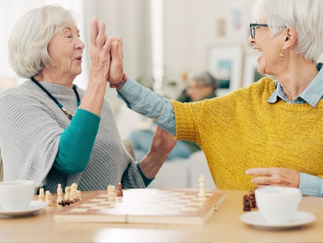 Two senior women celebrate with a high-five over a chessboard, smiling in a bright room with cups on the table.